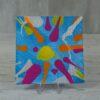 20260914_131706 Original 5x5 inch square abstract fluid painting on canvas panel featuring an explosive splash pattern of magenta, orange, lime green, and metallic silver paint droplets over a bright turquoise blue background, shown on a clear display stand.