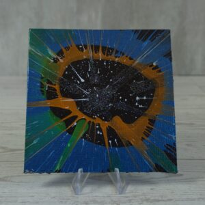 Original 5x5 inch square abstract spin painting on canvas panel featuring a dark black speckled central void surrounded by an orange-gold ring with sharp teal, green, and white paint radial streaks expanding outward across a dark blue canvas, shown on a clear plastic stand.