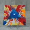 Original 5x5 inch square abstract spin painting on canvas panel featuring a central cobalt blue circular burst with sharp splash lines surrounded by circular streaks of red, orange, yellow, and blue paint radiating outward toward the canvas edges, displayed on a clear stand.